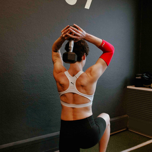 Sportlerin trainiert im Gym mit einer Hantel über dem Kopf und trägt eine rote neolevee Ellenbogenbandage.